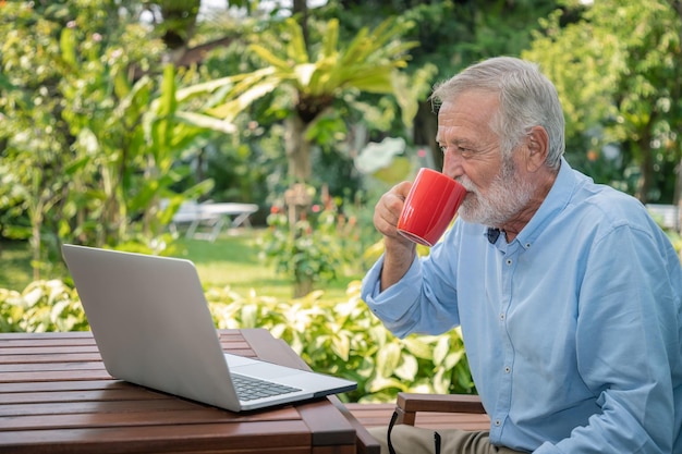 Leitender Mann mit weißen Haaren, der einen Computer-Laptop verwendet, der zu Hause arbeitet und Kaffee trinkt und eine Kamera sieht