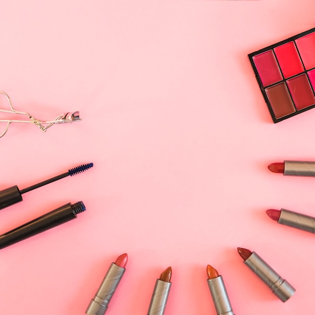 Lidschatten-Palette; Wimpernzange Mascara und verschiedene Schattierungen von Lippenstift über rosa Hintergrund