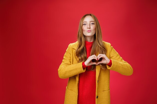 Liebe euch alle. Porträt einer romantischen und stilvollen, gut aussehenden, flirtenden Rothaarigen mit Sommersprossen und blauen Augen, die Lippen falten, um einen Kuss zu geben, der Herzgeste zeigt und in Sympathie über der roten Wand gesteht.