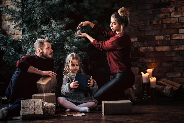 Liebe, Familie, Weihnachtszeit. Attraktive Familie schmückt den Weihnachtsbaum, umgeben von Geschenken zu Hause.