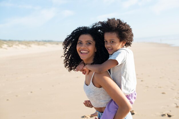 Liebende Mutter und Tochter verbringen Zeit am Strand. Afroamerikanische Familie geht spazieren, lacht, spielt, reitet auf dem Rücken. Freizeit, Familienzeit, Elternschaftskonzept