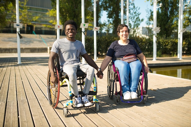 Liebender Mann und Frau im Rollstuhl verbringen Zeit in der Nähe von Wasser. Afroamerikanischer Mann und kaukasische Frau in Freizeitkleidung, Händchen haltend. Liebe, Zuneigung, Glückskonzept