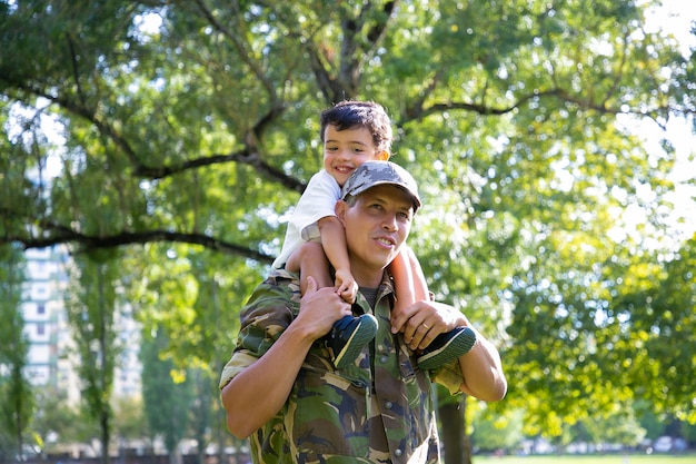 Liebender Vater, der Sohn am Hals hält und im Stadtpark geht. Glücklicher kaukasischer Sohn, der auf Hals des Vaters in der Uniform sitzt, ihn umarmt und wegschaut. Familientreffen, Vaterschaft und Rückkehr nach Hause Konzept