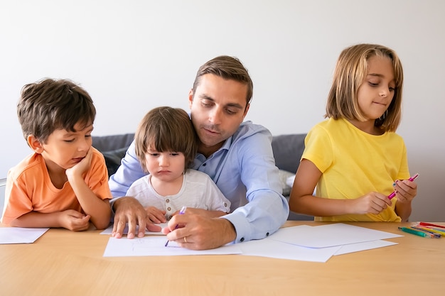 Liebender Vater und niedliche Kinder, die mit Markierung am Tisch zeichnen. Kaukasischer Vater mittleren Alters malt und spielt mit schönen Kindern im Wohnzimmer. Vaterschafts-, Kindheits- und Familienzeitkonzept