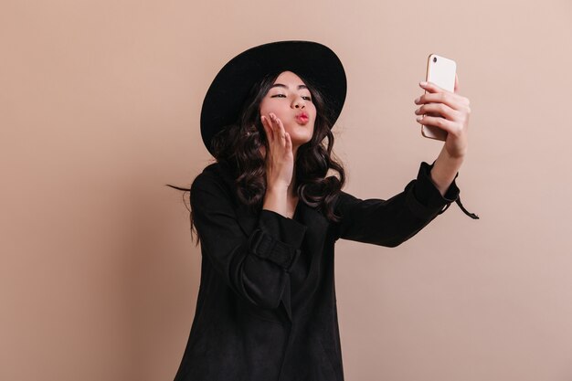 Liebenswerte chinesische Dame, die selfie nimmt. Winsome asiatische Frau mit Smartphone, das auf beigem Hintergrund aufwirft.