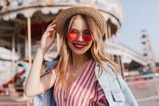 Liebenswerte junge Frau in der stilvollen rosa Sonnenbrille, die in guten Sommertag aufwirft. Außenporträt des romantischen weiblichen Modells, das im Vergnügungspark im Frühlingsmorgen kühlt.