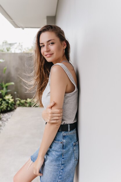 Liebenswertes weißes weibliches Modell im Jeansrock, der am Morgen lächelt. Außenfoto der attraktiven Frau mit dem langen braunen Haar, das nahe Wand aufwirft.