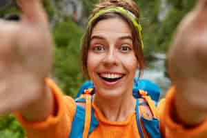 Kostenloses Foto liebevolle fröhliche touristin macht selfie-porträt, schaut glücklich in die kamera, steht mit rucksack gegen grünen naturraum