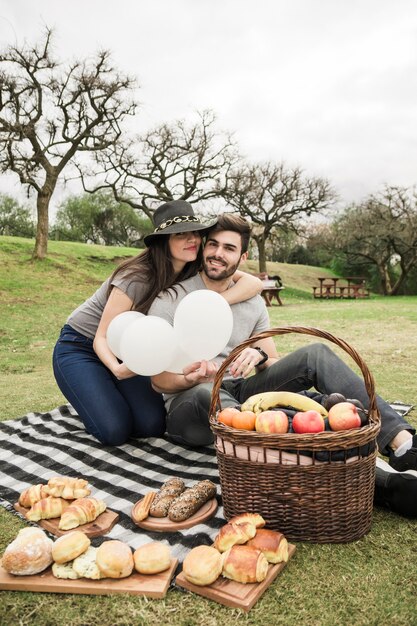 Liebevolle junge Paare, die mit gebackenem Lebensmittel und Früchten über dem Picknickkorb im Park sitzen