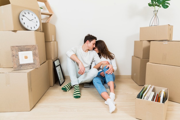 Liebevolle junge Paare, die zwischen dem Stapel Pappschachteln an ihrer neuen Wohnung sitzen