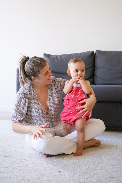 Liebevolle Mutter, die mit gekreuzten Beinen auf dem Boden sitzt und Baby ansieht. Nettes kleines Mädchen, das barfuß auf Teppich nahe lächelnder Mutter und beißende Hände steht. Familienzeit, Mutterschaft und Wochenendkonzept