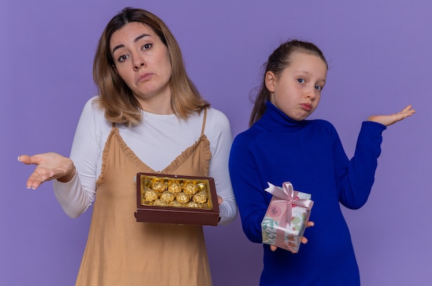 Liebevolle Mutter mit Schachtel Pralinen und Tochter, die Geschenk hält, das verwirrte Arme hebt, die internationalen Frauentag feiern, der über lila Wand steht