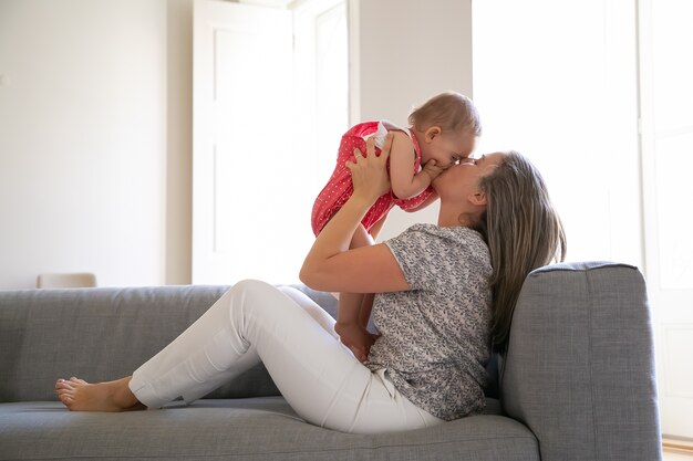 Liebevolle Mutter sitzt auf dem Sofa und küsst ihre kleine Tochter mit Liebe. Glückliches Baby, das Gesicht mit Handflächen schließt. Langhaarige Mutter hält Kind mit beiden Händen. Familien- und Mutterschaftskonzept