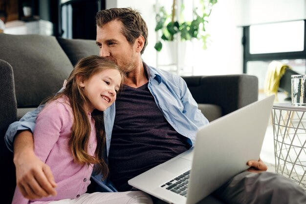 Liebevoller Vater, der seiner Tochter einen Kuss gibt, während er einen Computer benutzt und sich im Wohnzimmer entspannt