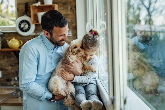 Liebevolles kleines mädchen, das einen hund umarmt, während es sich mit ihrem vater am fenster entspannt