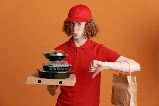 Lieferant Mitarbeiter in roter Kappe leere T-Shirt Uniform hält Pizzakarton Lebensmittelbehälter mit Papiertüte, die unzufrieden in die Kamera schaut und einen schiefen Mund macht, der den Daumen nach unten zeigt, der über orangefarbenem Hintergrund steht