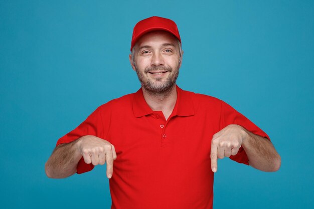 Lieferant Mitarbeiter in roter Kappe leere T-Shirt Uniform zeigt mit Zeigefinger nach unten und lächelt selbstbewusst in die Kamera, die über blauem Hintergrund steht