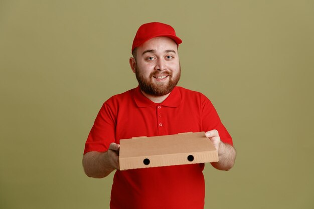 Lieferbote-Angestellter in der roten Kappen-T-Shirt-Uniform, die den Pizzakarton hält, der glücklich und positiv lächelnd in die Kamera blickt und fröhlich über grünem Hintergrund steht