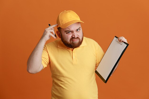 Lieferbote-Angestellter in gelber Kappe, leerem T-Shirt, Uniform, Klemmbrett und Stift, der verwirrt und unzufrieden in die Kamera blickt und auf seinen Kopf zeigt, der über orangefarbenem Hintergrund steht