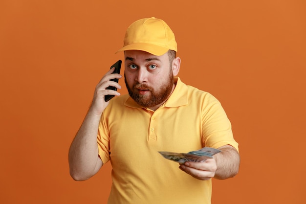 Lieferbote-Angestellter in gelber Kappen-T-Shirt-Uniform, die Bargeld hält und am Handy spricht, glücklich und positiv über orangefarbenem Hintergrund