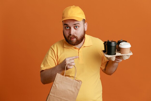 Lieferbote-Angestellter in gelber Kappen-T-Shirt-Uniform mit Kaffeetassen und Papiertüten, die mit dem Zeigefinger auf Tassen zeigen und überrascht aussehen, wie sie über orangefarbenem Hintergrund stehen