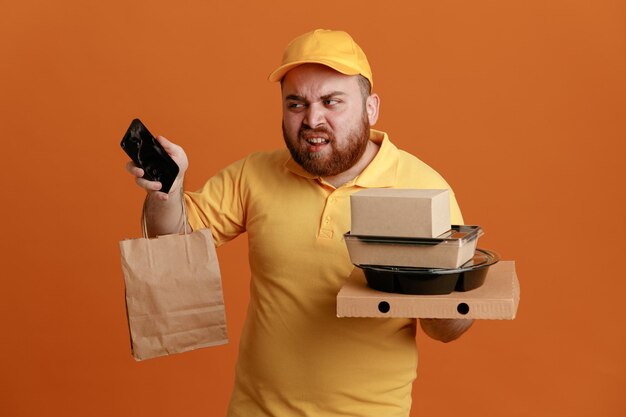 Lieferbote-Angestellter in gelber Kappen-T-Shirt-Uniform mit Lebensmittelbehälter und Pizzaschachtel mit Papiertüte, die das Smartphone hält und verwirrt über orangefarbenem Hintergrund steht