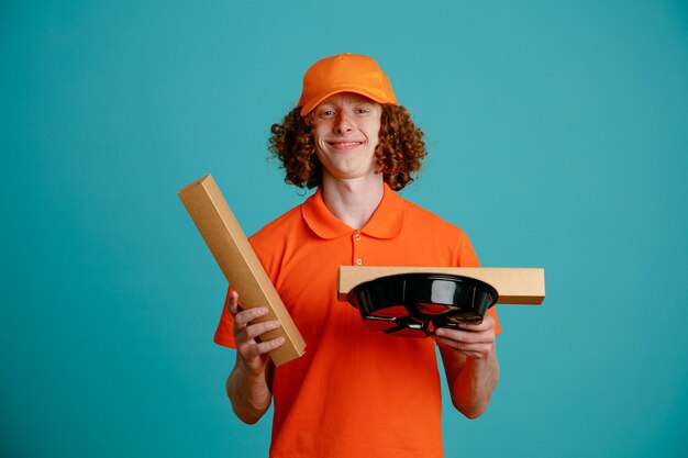 Lieferbote-Angestellter in orangefarbener T-Shirt-Uniform, die Pizzakarton und Lebensmittelbehälter hält und in die Kamera schaut, glücklich und positiv lächelnd, selbstbewusst über blauem Hintergrund