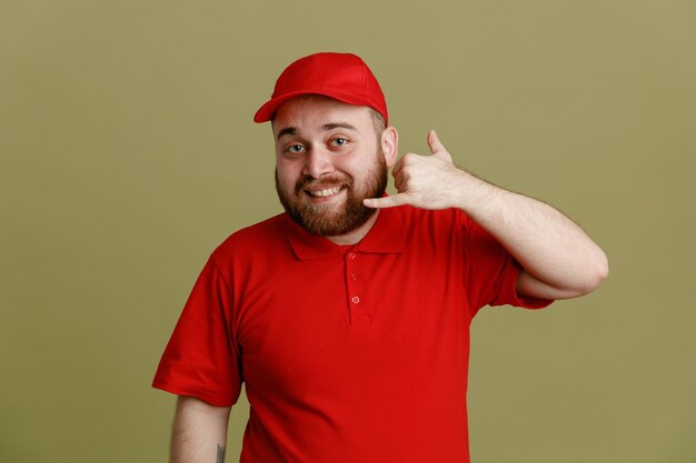 Lieferbote-Angestellter in roter Kappe, leerem T-Shirt, einheitlichem Blick in die Kamera, glücklich und positiv lächelnd, fröhlich machend, rufen Sie mich an, Geste mit der Hand, die über grünem Hintergrund steht