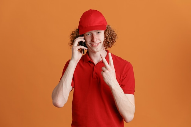 Lieferbote-Angestellter in roter Kappe, leerem T-Shirt, Uniform, der auf dem Handy spricht und lächelnd in die Kamera schaut und die Nummer zwei oder Vsign zeigt, die über orangefarbenem Hintergrund stehen