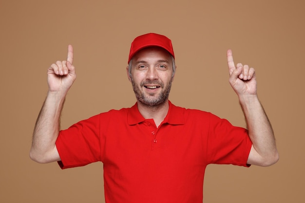 Kostenloses Foto lieferbote-angestellter in roter kappe, leerem t-shirt, uniform, der mit den zeigefingern nach oben zeigt und selbstbewusst in die kamera schaut, die über braunem hintergrund steht