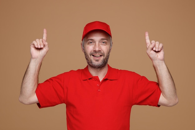 Lieferbote-Angestellter in roter Kappe, leerem T-Shirt, Uniform, der mit den Zeigefingern nach oben zeigt und selbstbewusst in die Kamera schaut, die über braunem Hintergrund steht