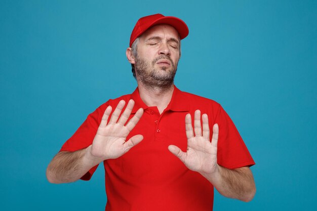 Lieferbote-Angestellter in roter Kappe, leerem T-Shirt, Uniform, der unzufrieden aussieht und eine Verteidigungsgeste macht, die sich weigert, über blauem Hintergrund zu stehen