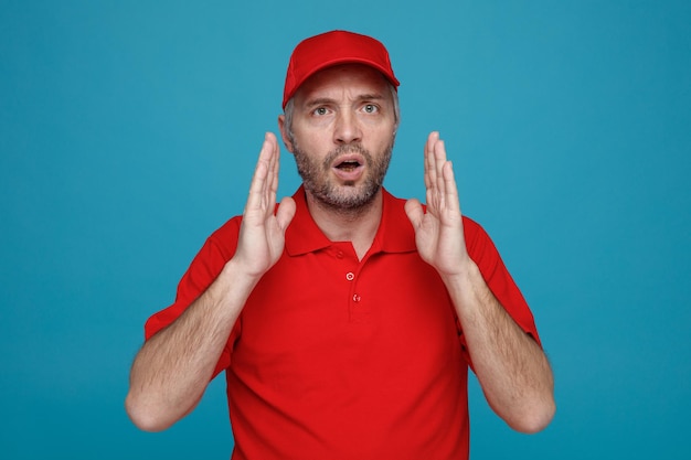 Lieferbote-Angestellter in roter Kappe, leerem T-Shirt, Uniform, der verwirrt in die Kamera blickt und mit den Händen, die über blauem Hintergrund stehen, eine Größengeste macht