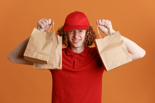 Lieferbote-Angestellter in roter Kappe, leerem T-Shirt, Uniform, die Papiertüten hält, die in die Kamera schauen, glücklich und positiv lächelnd, selbstbewusst über orangefarbenem Hintergrund