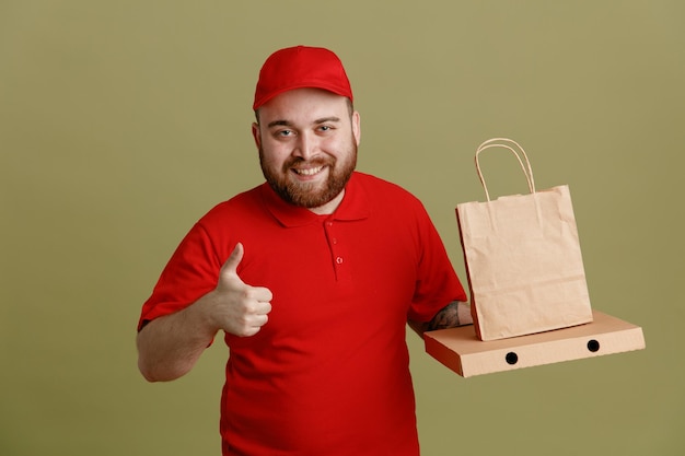 Lieferbote-Angestellter in roter Kappe, leerem T-Shirt, Uniform mit Pizzaschachtel und Papiertüte, die in die Kamera blickt, glücklich und positiv lächelnd und fröhlich zeigt, Daumen nach oben stehend über grünem Hintergrund