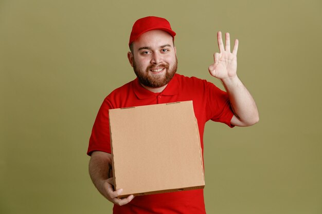 Lieferbote-Angestellter in roter Kappe, leerem T-Shirt, Uniform, Pizzakarton haltend, der in die Kamera blickt, glücklich und positiv lächelnd und fröhlich zeigt, ok-Zeichen, das über grünem Hintergrund steht