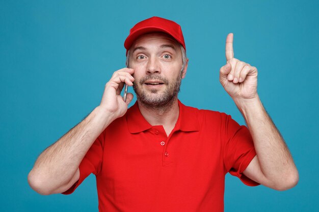 Lieferbote-Angestellter in roter Kappe, leerer T-Shirt-Uniform, der am Handy lächelt, glücklich und positiv zeigt und den Zeigefinger zeigt, der eine gute Idee hat, über blauem Hintergrund zu stehen