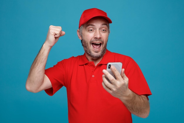 Lieferbote-Angestellter in roter Kappe, leerer T-Shirt-Uniform, der das Smartphone hält, das auf den Bildschirm schaut, verrückt, glücklich und aufgeregt, die Faust ballt und sich über seinen Erfolg freut, der über blauem Hintergrund steht