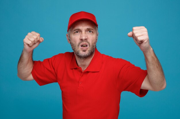 Lieferbote-Angestellter in roter Kappe, leerer T-Shirt-Uniform, der in die Kamera schaut, mit wütendem Gesicht, das die Fäuste ballt und bereit ist, über blauem Hintergrund zu kämpfen