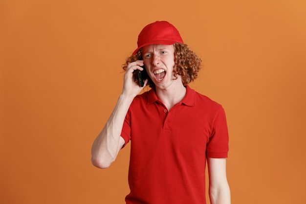 Lieferbote-angestellter in roter kappe, leerer t-shirt-uniform, der wütend und enttäuscht auf dem handy spricht und über orangefarbenem hintergrund schreit und schreit Premium Fotos