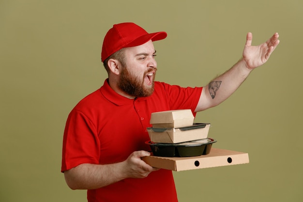 Kostenloses Foto lieferbote-angestellter in roter kappe, leerer t-shirt-uniform, die lebensmittelbehälter und pizzakarton hält und beiseite schaut, wütend und frustriert schreit und den arm in unmut hebt, der über grünem hintergrund steht