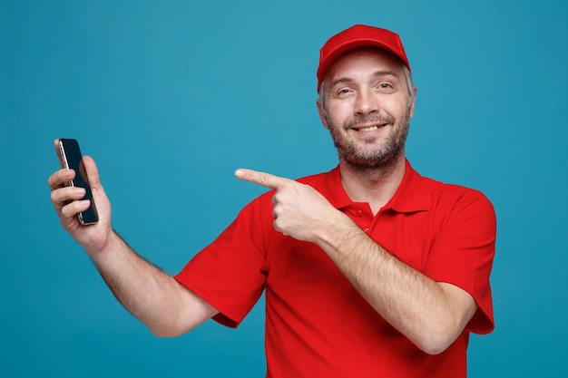 Lieferbote-Angestellter in roter Kappe, leeres T-Shirt, einheitliches Smartphone, das mit dem Zeigefinger darauf zeigt und mit einem Lächeln auf dem glücklichen Gesicht, das über blauem Hintergrund steht, in die Kamera blickt