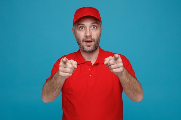 Lieferbote-Angestellter in roter Kappe, T-Shirt-Uniform, der erstaunt und überrascht in die Kamera blickt und mit beiden Händen auf die Kamera zeigt, die über blauem Hintergrund steht