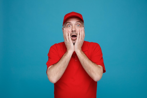Lieferbote-Angestellter in roter Kappen-T-Shirt-Uniform, der erstaunt und überrascht in die Kamera blickt und den Mund bedeckt, während die Hände über blauem Hintergrund stehen