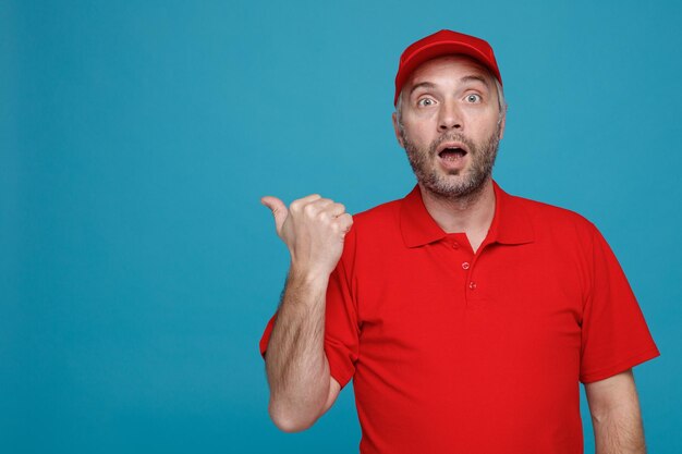 Lieferbote-Angestellter in roter Kappen-T-Shirt-Uniform, der erstaunt und überrascht in die Kamera blickt und mit dem Daumen auf die Seite zeigt, die über blauem Hintergrund steht