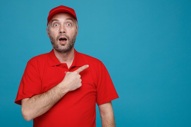Lieferbote-Angestellter in roter Kappen-T-Shirt-Uniform, der erstaunt und überrascht in die Kamera blickt und mit dem Zeigefinger auf die Seite zeigt, die über blauem Hintergrund steht