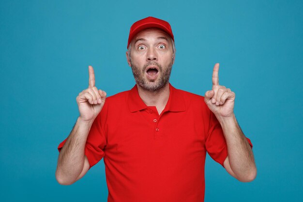 Lieferbote-Angestellter in roter Kappen-T-Shirt-Uniform, der erstaunt und überrascht in die Kamera blickt und mit den Zeigefingern nach oben zeigt, wobei beide Hände über blauem Hintergrund stehen