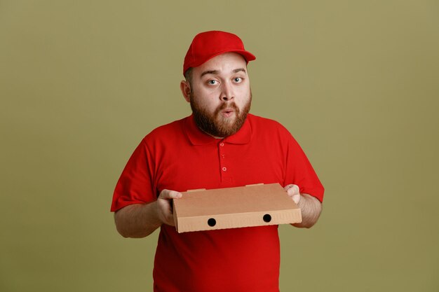 Lieferbote-Angestellter in roter Kappen-T-Shirt-Uniform mit Pizzakarton, der glücklich und überrascht in die Kamera blickt und über grünem Hintergrund steht