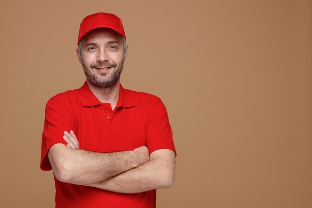 Lieferbote-Angestellter in roter T-Shirt-Uniform, die mit verschränkten Armen in die Kamera blickt und selbstbewusst, glücklich und positiv über braunem Hintergrund steht