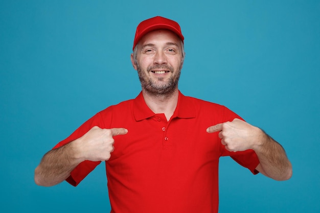 Lieferbote-Angestellter in roter T-Shirt-Uniform mit leeren Kappen, der mit Zeigefingern auf sich selbst zeigt und selbstbewusst lächelt, während er glücklich und erfreut über blauem Hintergrund in die Kamera blickt